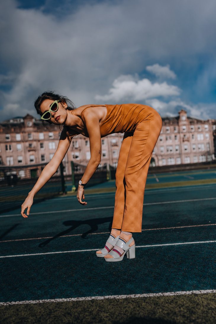 Leaning forward in an orange jumpsuit and sunglasses on a rooftop court